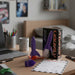 A purple rocket from heebie Jeebies and orange rocket-shaped personal massager displayed on a wooden desk next to its black and orange retail box, with a laptop, mug, and bedroom background visible.